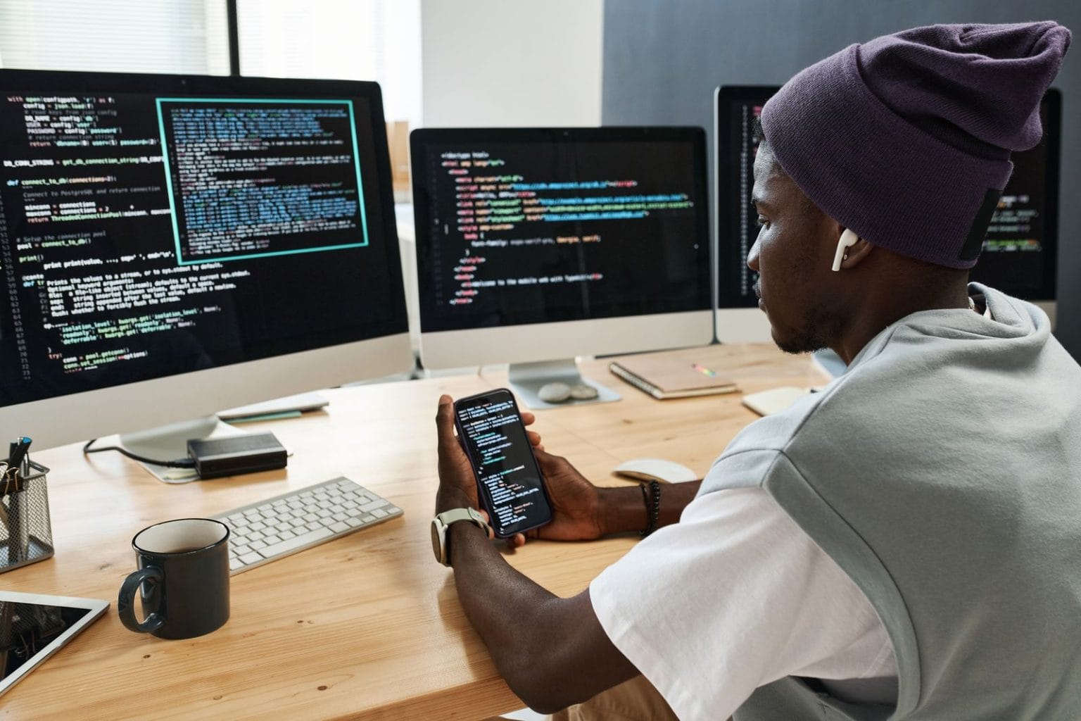 Developer testing a mobile app on a phone next to monitors showing code