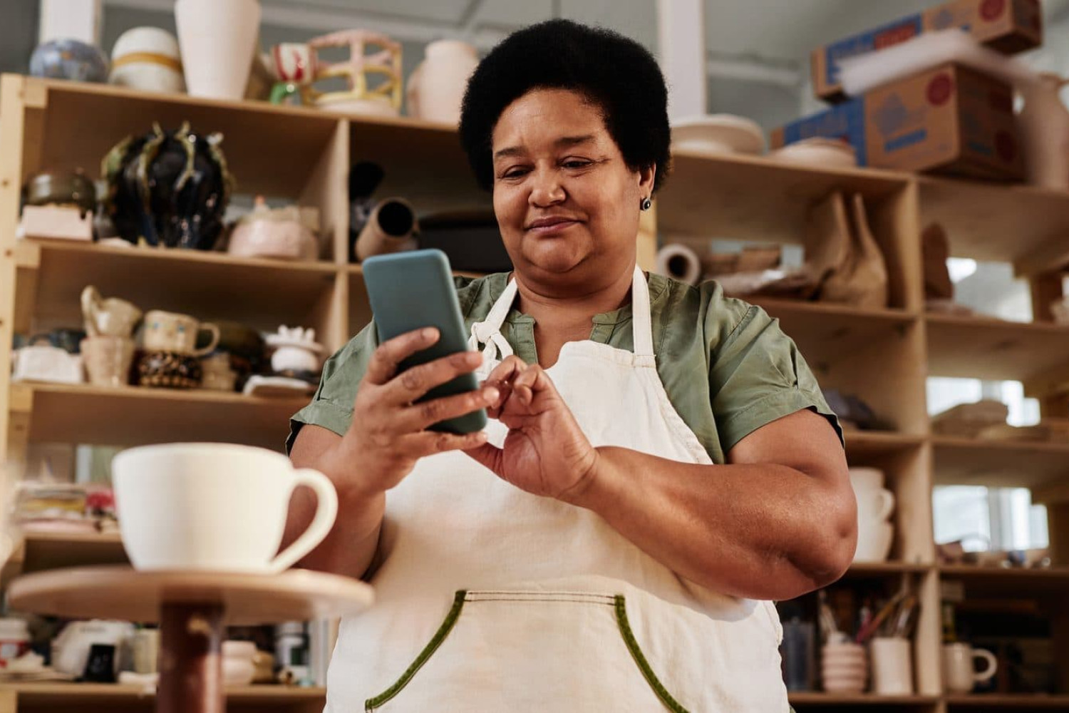 Small business owner in an apron checking her phone in her pottery shop