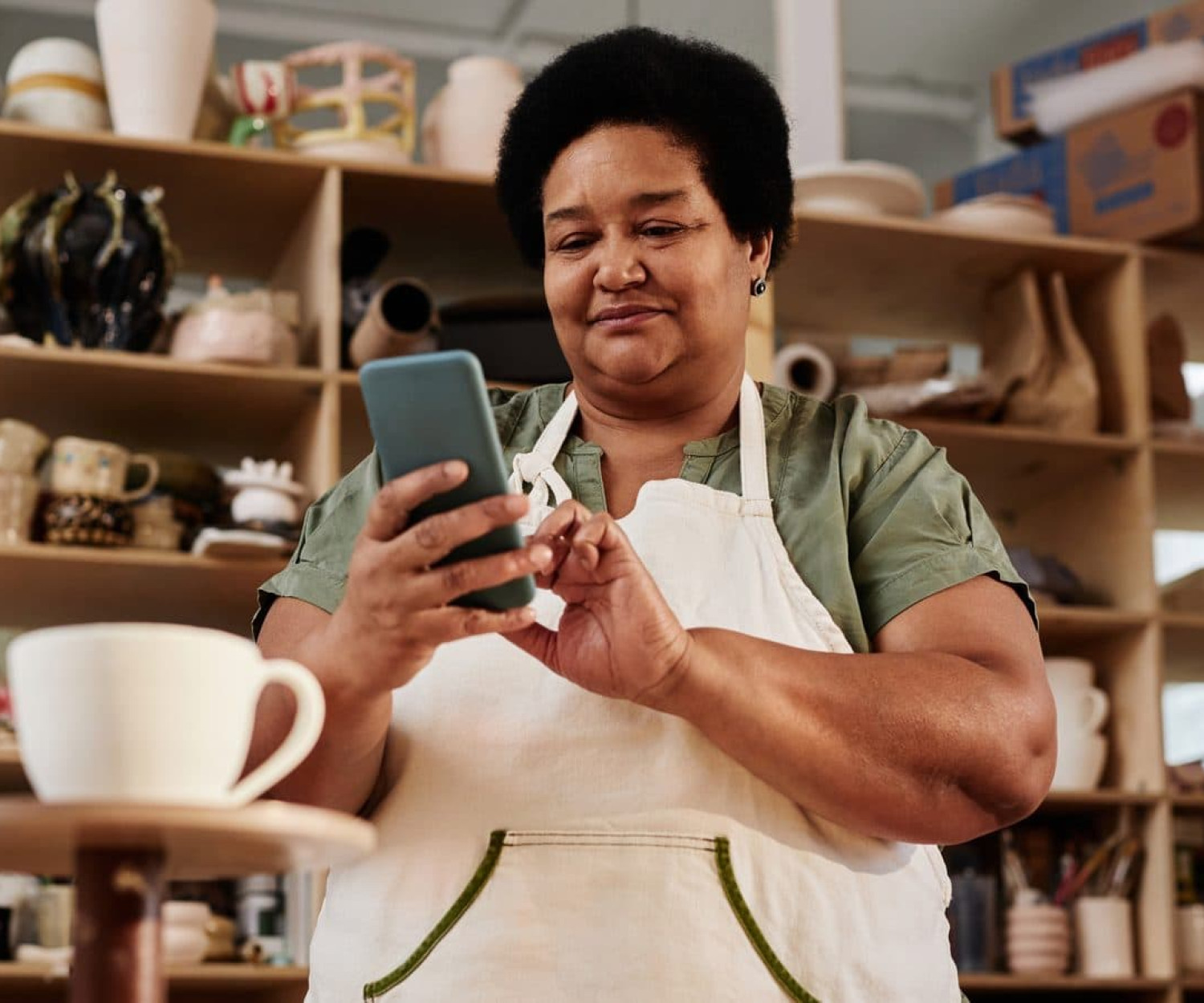 Small business owner in an apron checking her phone in her pottery shop