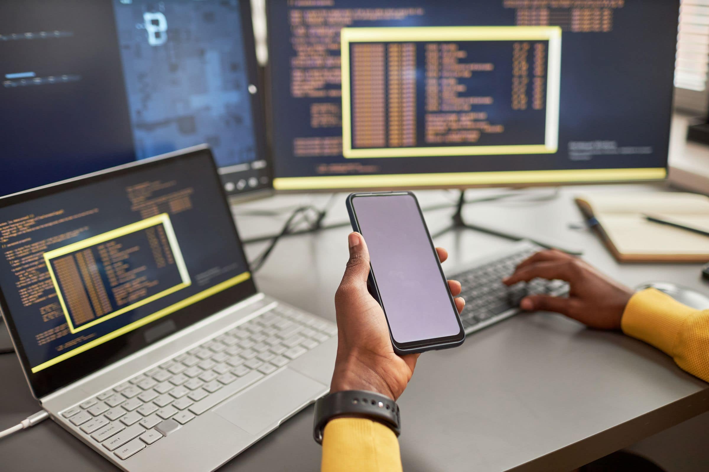 Developer holding a phone in front of a laptop and monitor showing code